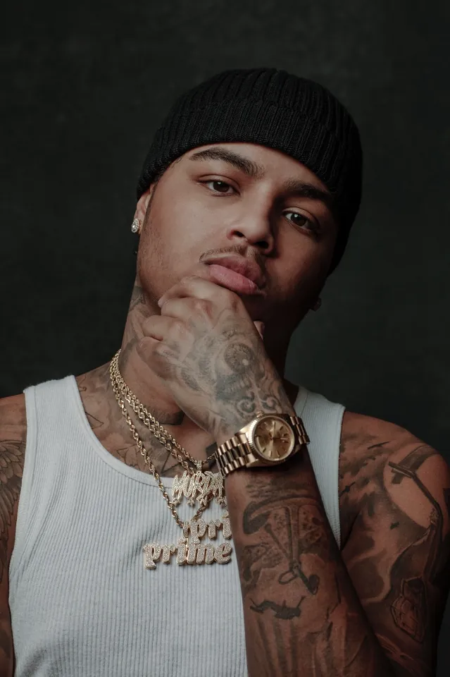 Jay Whiss in a black beanie and white tank top, hand to chin, wearing gold chains and a gold watch