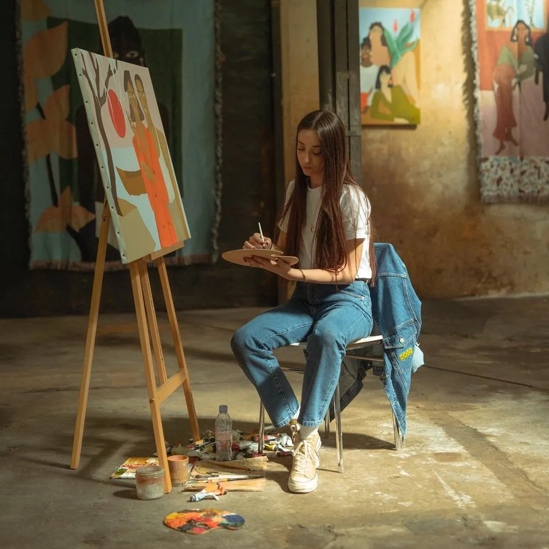 Léa Augereau at an easel in a concrete-floored studio, palette in hand, her paintings of women on the walls behind